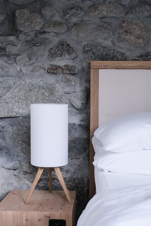 Un letto con lenzuola bianche, un muro in pietra, un comodino con una lampada e un portaoggetti in legno.