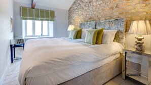 Quarto com parede de pedra, cama com roupa de cama branca, escrivaninha com cadeira e luminária.