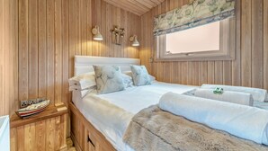 Une pièce aux murs lambrés, comprenant un lit, une table de chevet, une fenêtre ornée d’un rideau à motifs floraux et des lampes fixées au mur.