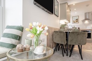 Una sala de estar moderna con un sofá, una mesa de centro de vidrio, un florero con flores y una cocina al fondo.