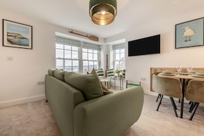 Una sala de estar con un sofá verde, una mesa de comedor con sillas, un televisor montado en la pared y una ventana con cortinas.