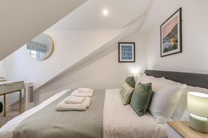 Un dormitorio con cama, almohadas, una mesita de noche, lámpara, espejo y dos cuadros en la pared.
