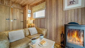 A cozy room with wooden walls, a sofa, a table with a plate of food and cups, and a wood-burning stove. - 1 Bed in Gatehouse Of Fleet (oc-d34894) (Gatehouse of Fleet)