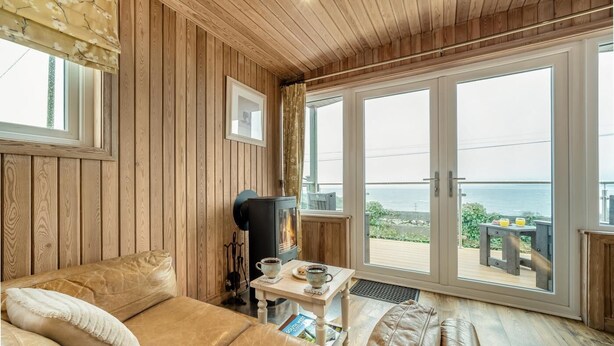 A cozy room with a wooden interior, a sofa, a table with a cup and a book, and a view of the sea through large glass doors. - 1 Bed in Gatehouse Of Fleet (oc-d34894) (Gatehouse of Fleet)