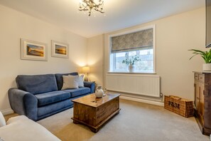 A living room with a blue sofa, a wooden coffee table, a lamp, a window with a view, and a radiator. - 2 Bed in Dersingham (oc-t35169) (Dersingham)