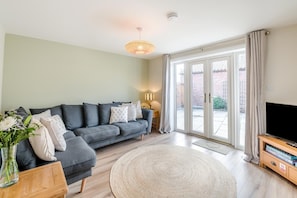 Ein Wohnzimmer mit grauem Sofa, einem runden Teppich, einer TV-Konsole mit Büchern und einer Glastür zu einem Außenbereich.