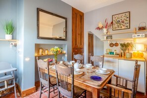 A dining area with a wooden table set for four, surrounded by chairs. A mirror hangs on the wall above the table, and there are shelves with decorative items and a potted plant to the left. - 2 Bed in Conwy (oc-t36008) (Conwy)
