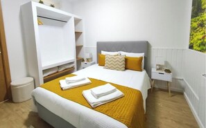 A neatly made bed with a white and yellow bedspread, a grey headboard, and two pillows. There is a white wardrobe with shelves, a small bedside table with a lamp, and a white nightstand with a lamp.
