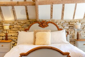 Ein Bett mit Holzvorhang, weißer Bettwäsche und einem flauschigen Kissen. Zwei Nachttische mit Lampen stehen bereit. Der Raum hat eine Steinwand und Holzbalken an der Decke.