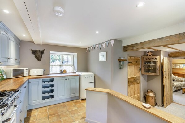 A kitchen with a stove, microwave, and white refrigerator. There is a window with curtains and a bull head decoration on the wall. - 2 Bed in Burton Bradstock (oc-o35069) (Burton Bradstock)