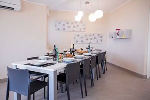 Un espace repas avec une longue table dressée pour accueillir plusieurs personnes, des chaises, un climatiseur fixé au mur et deux luminaires fixés au mur.