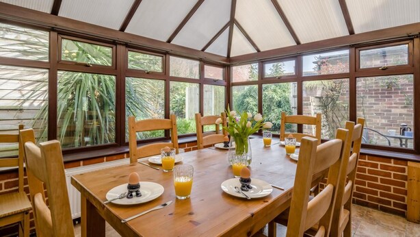 Ein Wintergarten mit einem Holztisch, gedeckt für das Frühstück, umgeben von Stühlen und großen Fenstern mit Blick auf viel Grün.
