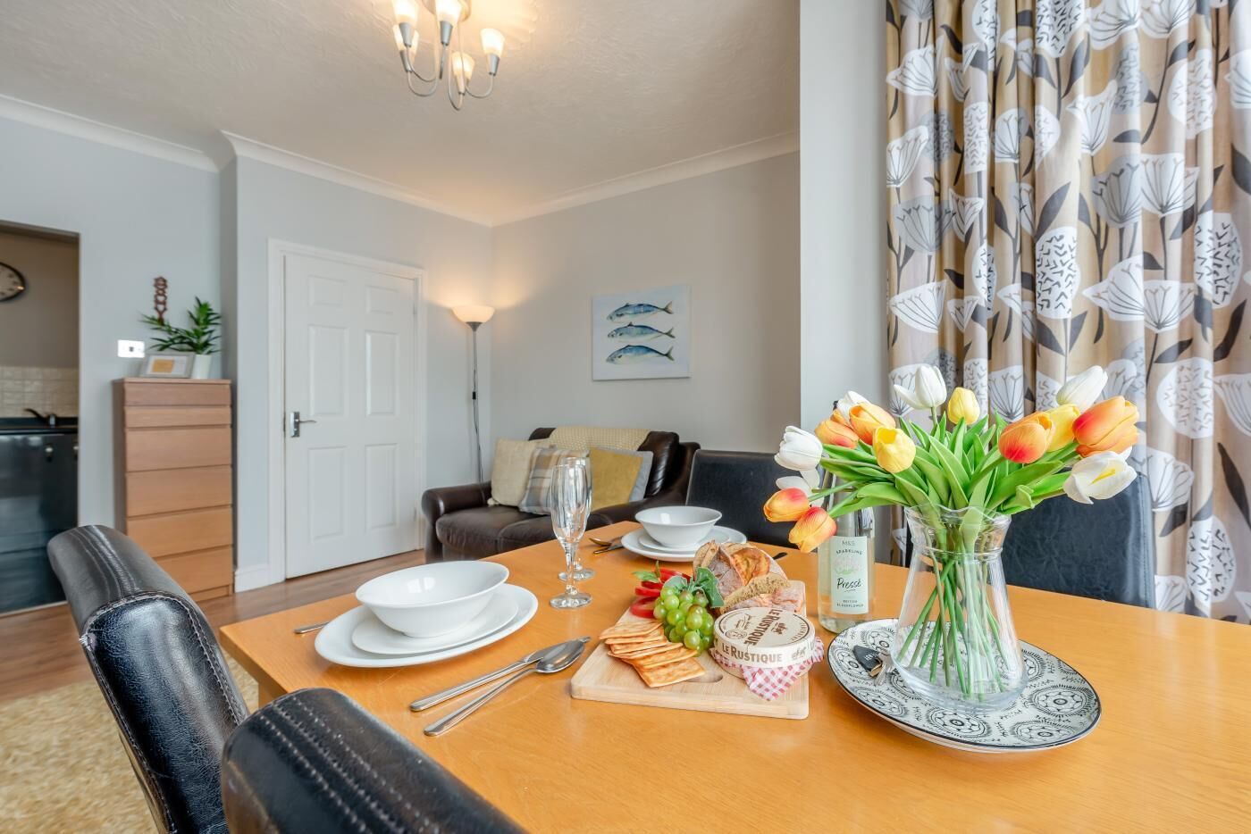 A dining area with a wooden table set for two, a vase of tulips, and a bottle of wine.