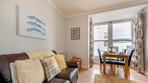 A living room with a brown leather sofa, a dining table with chairs, a rug, a painting of fish, and a framed picture on the wall. - 1 Bed in Tenby (oc-t36054) (Tenby)
