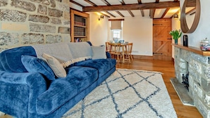 A cozy living room with a blue sofa, a stone wall, wooden beams, a dining area, and a fireplace. - 2 Bed in Whitby (oc-dedi4) (Whitby)