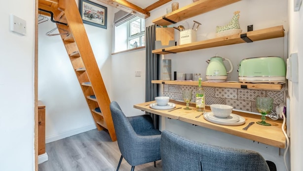 A compact kitchen with wooden shelves, a dining table, and two chairs. - 2 Bed in Whitby (oc-dedi4) (Whitby)