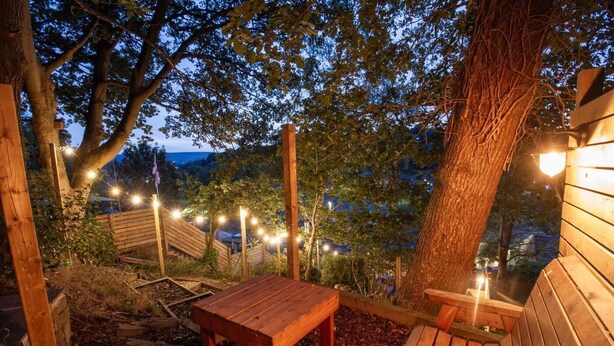 Ein Sitzbereich im Freien mit Holzbänken und einem Tisch, umgeben von Bäumen und Lichterketten.