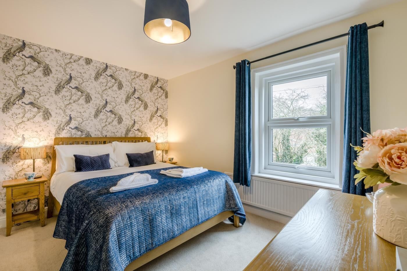 A bedroom with a bed, bedside lamps, a window with curtains, and a vase with flowers.