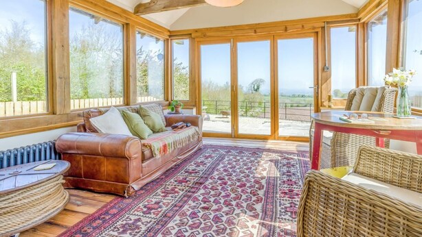 A sunlit room with a leather sofa, a patterned rug, and a view through large glass doors to an outdoor area. - 1 Bed in Picklescott (oc-o35147) (Ratlinghope)