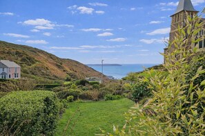 A coastal landscape with a grassy area, a stone tower, and a building with a pitched roof. - 4 Bed in Mortehoe (oc-d35057) (Mortehoe)