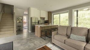 A modern kitchen with a large island, a built-in oven, and a refrigerator. There is a staircase leading to another level, a sofa with a cushion, and a window with a view of greenery. - Forest immersion | 5 min beach • 15 min Tremblant (Les Laurentides Regional County Municipality)