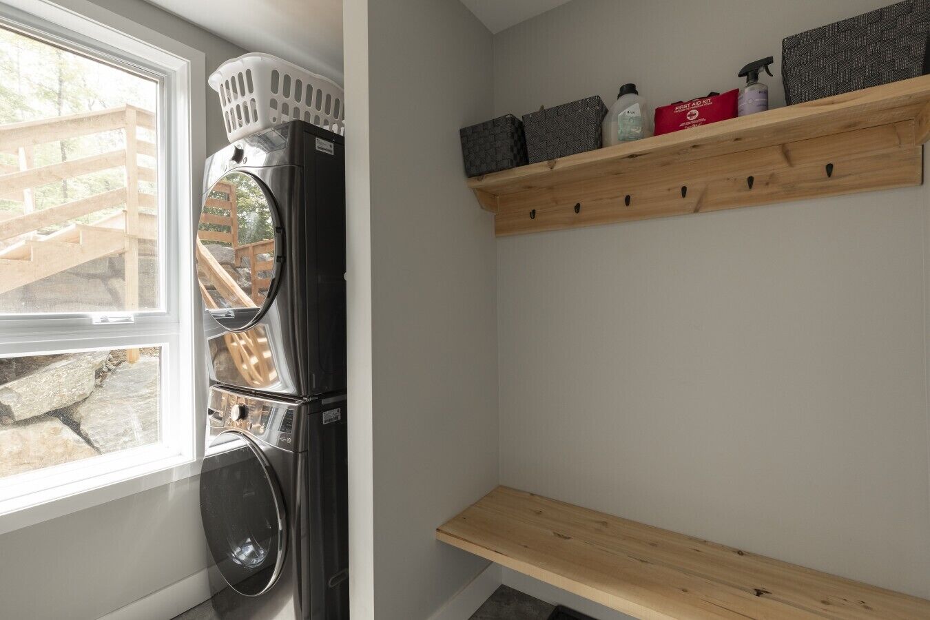 A laundry area with a stacked washer and dryer, a wooden shelf with storage boxes, and a window with a view of a wooden deck.