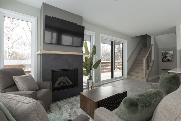 A modern living room with a fireplace, large windows, and a staircase.