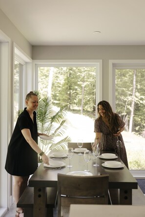 A dining area with a table set for two, large windows, and a view of greenery outside.
