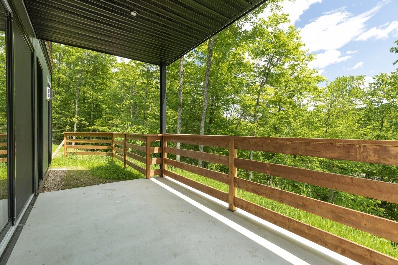 Een overdekt buitengebied met houten balustrade, betonnen vloer en uitzicht op een bebost gebied.