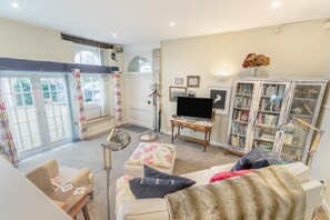 A living room with a sofa, a television, a bookshelf, and a large window with curtains. - 1 Bed in Fowey (oc-t36056) (Fowey)