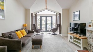 A modern living room with a grey sofa, ottoman, and two armchairs, a flat-screen TV, and a fireplace. - 4 Bed in Brixham (oc-t35056) (Brixham)