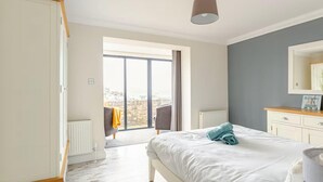 A bedroom with a large bed, a dresser, a chair, and a view of the city through a large window. - 4 Bed in Brixham (oc-t35056) (Brixham)
