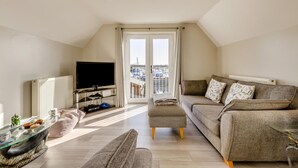 Un salón amplio con televisor, sofá y reposapiés, una mesa de centro de vidrio y vistas de la ciudad a través de amplias ventanas.