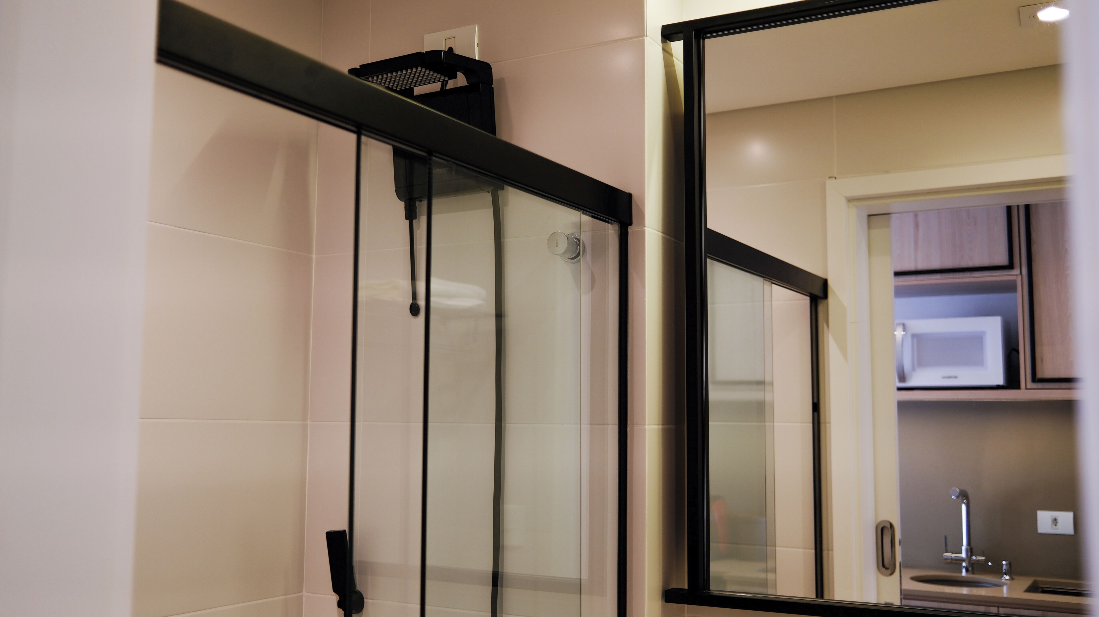 A modern bathroom with a glass shower enclosure, a black towel rack, and a visible microwave in the adjacent kitchen area.