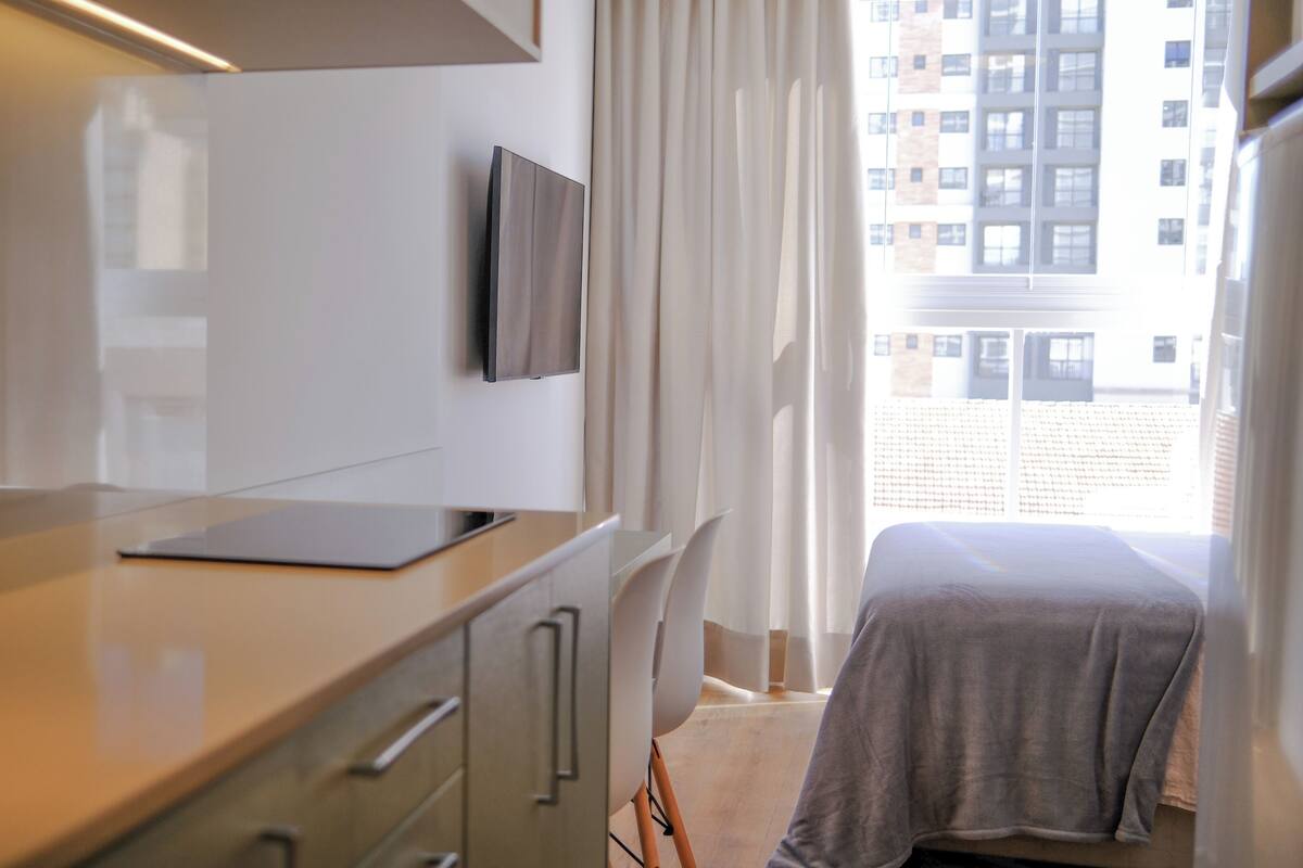 A modern hotel room with a flat-screen TV mounted on the wall, a small desk with a laptop, a bed with a grey blanket, and a view of a cityscape through the window.