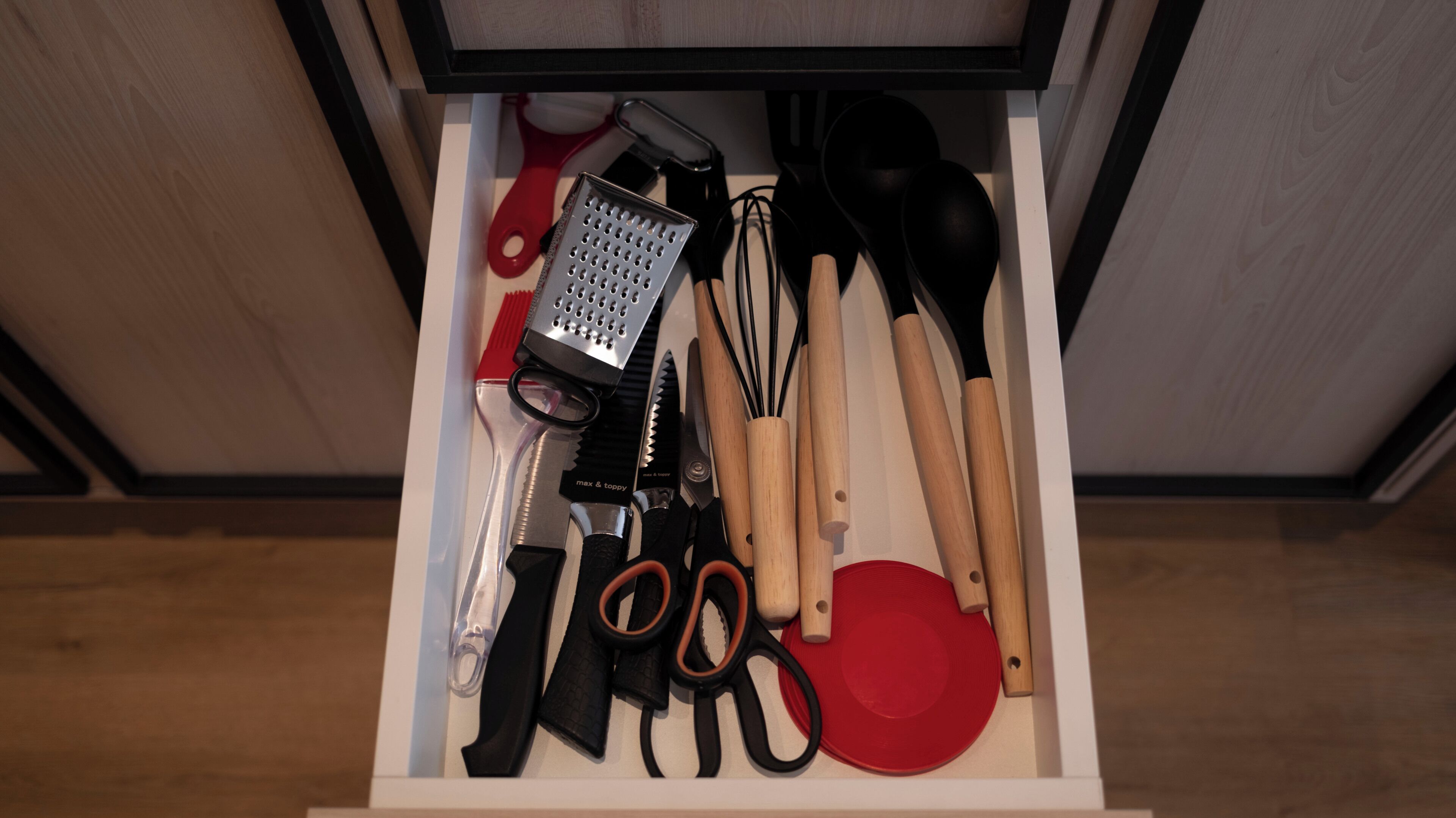 A drawer with kitchen utensils including a grater, whisk, spatula, and a red plate.