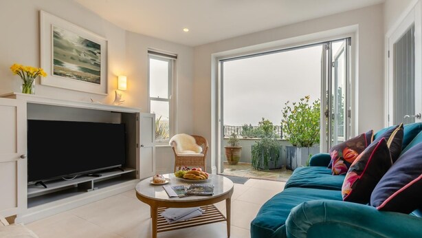 Un salon moderne avec un téléviseur à écran plat, un fauteuil en osier, un canapé de couleur turquoise et un balcon agrémenté de plantes en pot.
