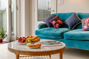 Ein runder Holztisch mit einer Fruchtschale, einer Donutplatte und einer Zeitschrift. Im Hintergrund steht ein türkisfarbenes Sofa mit floralen Kissen.
