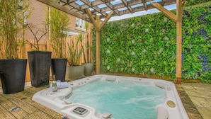 Un jacuzzi en un área exterior cubierta con una pérgola de madera, rodeado de plantas en macetas y un muro de ladrillo.