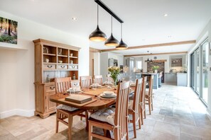 Een eetgedeelte met een houten tafel voor zes personen, omringd door stoelen. Achtergrond is een keuken met een groot kookeiland, en de kamer heeft een hoge plafond met hanglampen.