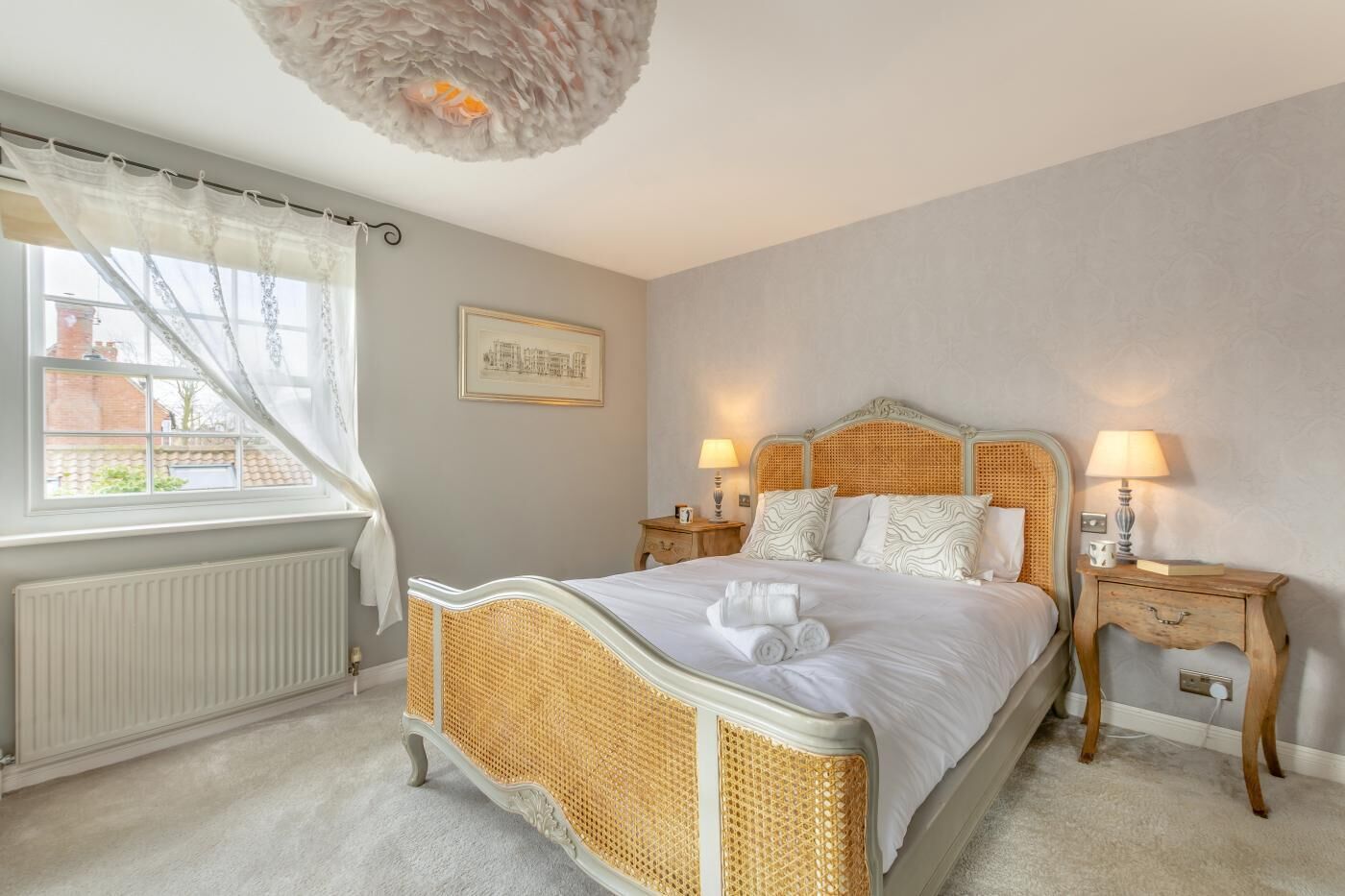 Quarto com cama, mesas de cabeceira, janela com cortinas, radiador e um quadro na parede.