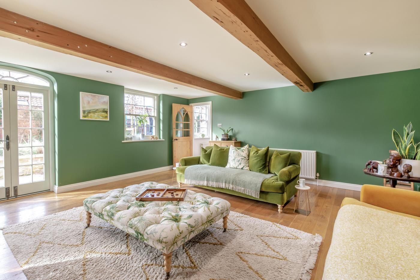 Ein Zimmer mit grüner Wand, Holzbalken, einem grünen Sofa, einem gemusterten Hocker und einem gelben Sessel.