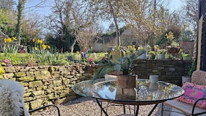A round glass table with wicker chairs, a stone wall, and a variety of potted plants and flowers. - 3 Bed in Ellwood (oc-t35963) (Ellwood)