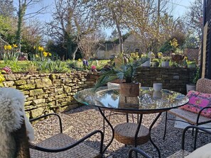 A round glass table with wicker chairs, a stone wall, and a variety of potted plants and flowers. - 3 Bed in Ellwood (oc-t35963) (Ellwood)