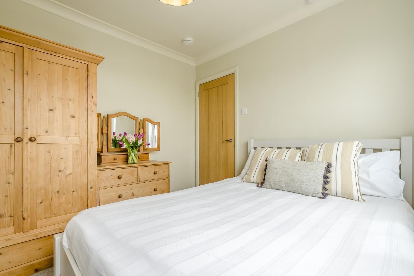 Een slaapkamer met een bed, een houten kledingkast, een dressoir met spiegel en een deur.