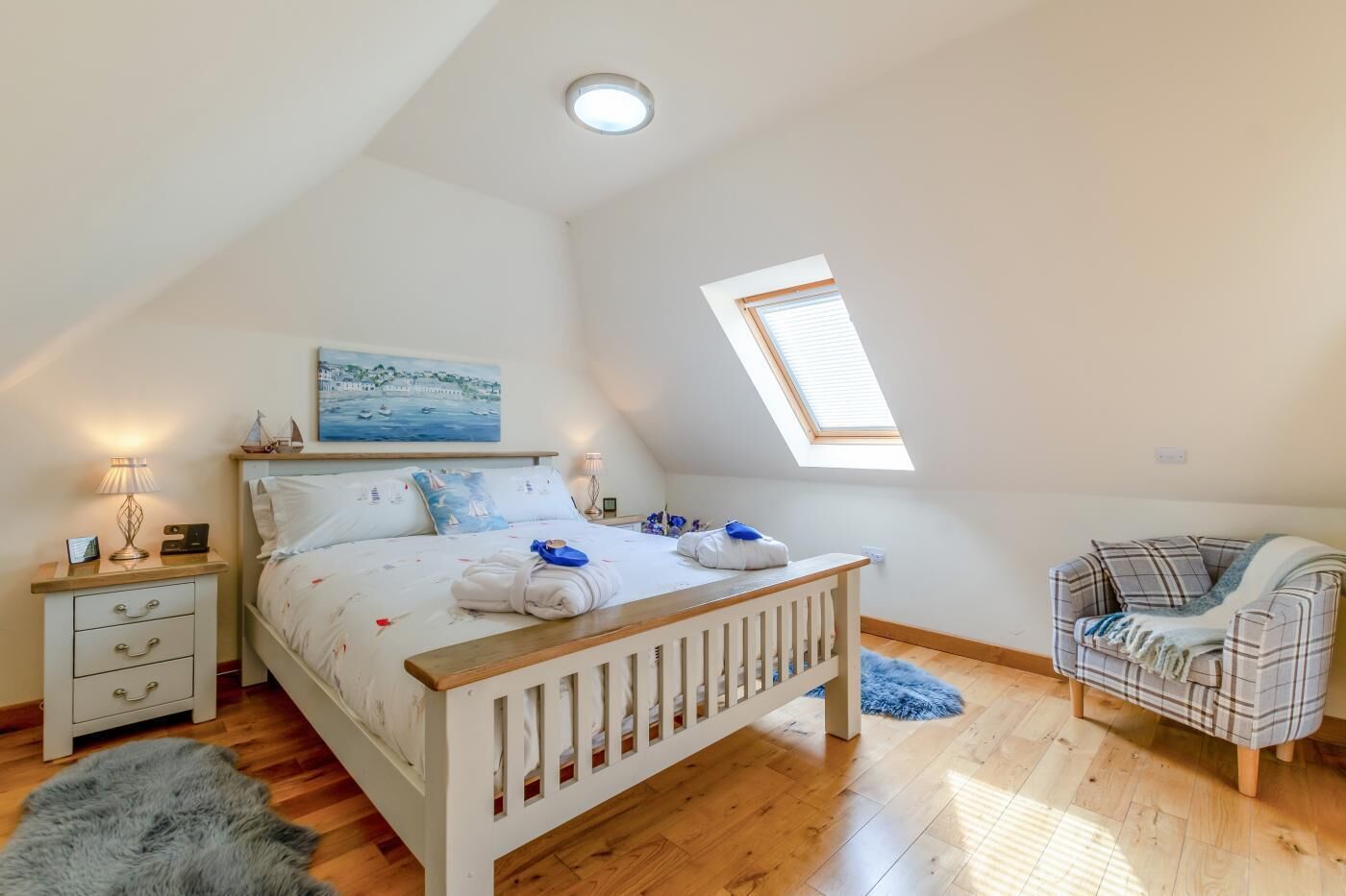 Ein Schlafzimmer mit schrägen Wänden, einem Oberlicht, einem Bett mit Kopfteil, einem Nachttisch mit Lampe, einem Sessel und einem Teppich.