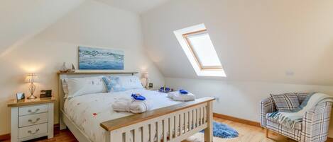 Ein Schlafzimmer mit schrägen Wänden, einem Oberlicht, einem Bett mit Kopfteil, einem Nachttisch mit Lampe, einem Sessel und einem Teppich.