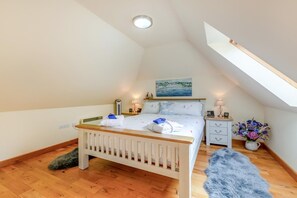 A bedroom with a sloped ceiling, a bed, bedside tables, a nightstand, a painting, a vase with flowers, and a fluffy rug.
