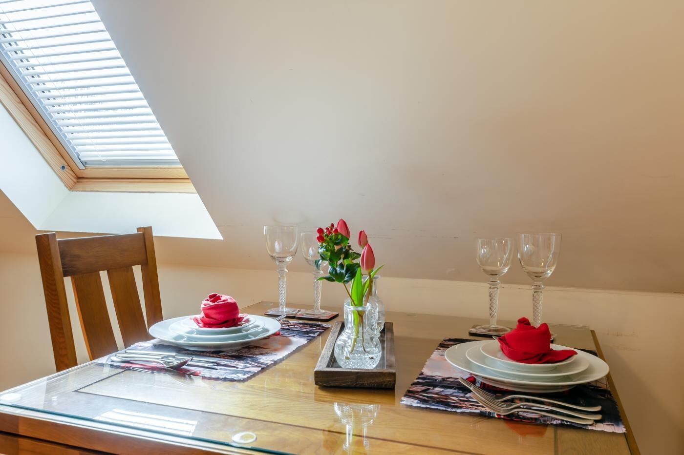 Een eettafel met een glazen blad, twee stoelen, een vaas met bloemen en wijnglazen.