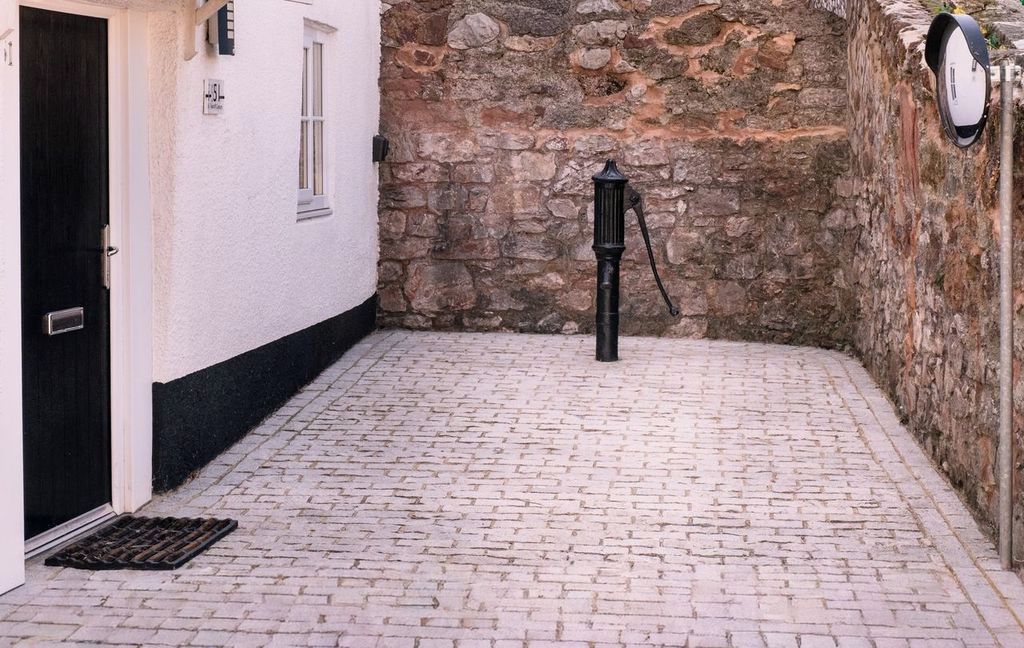 A narrow cobblestone alleyway with a stone wall on one side and a white building with a black door on the other. A black lantern is mounted on the wall.